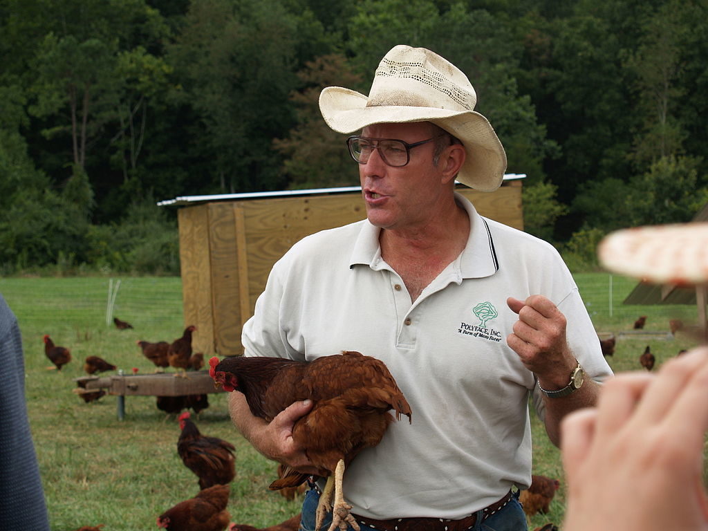 Man in cowboy hat holding rust colored chicken, gesturing with his hands